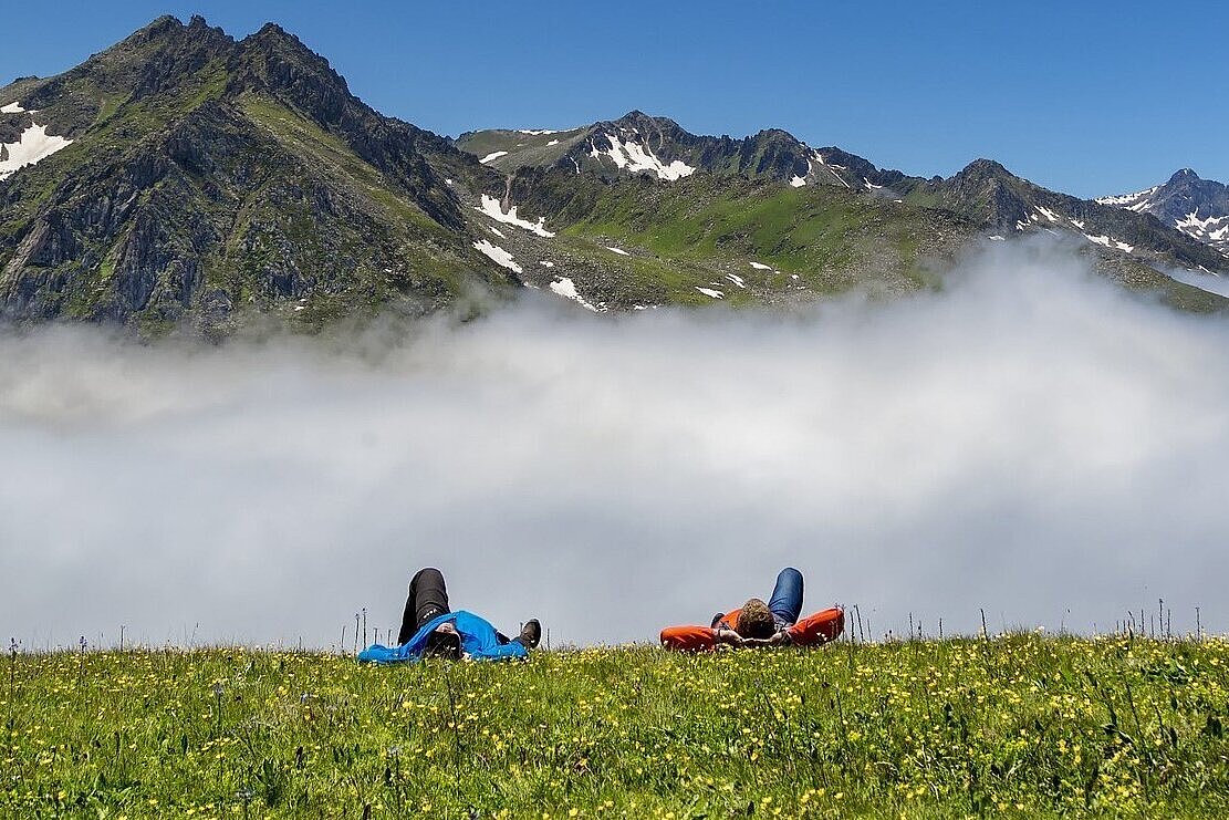 Menschen liegen auf der Wiese vor einem Gebirgsmassiv unter blauem Himmel.