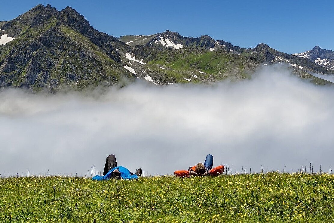 Menschen liegen auf der Wiese vor einem Gebirgsmassiv unter blauem Himmel.
