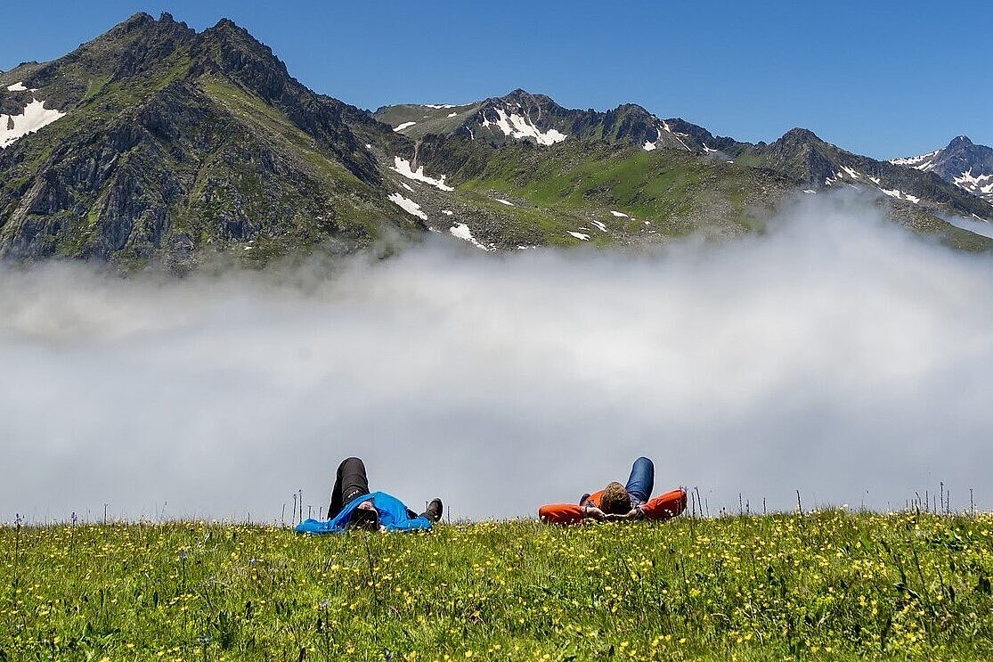 Menschen liegen auf der Wiese vor einem Gebirgsmassiv unter blauem Himmel.