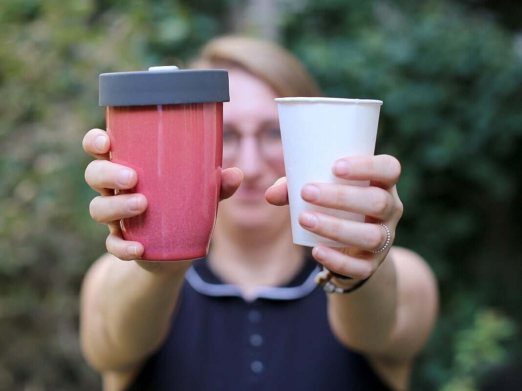Frau hält zwei Becher. Einen roten Mehrwegbecher und einen weißen Einweg-Plastikbecher.