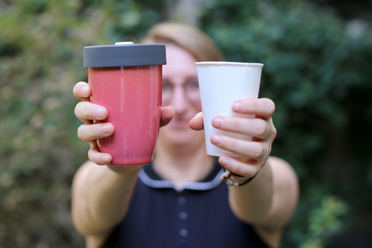 Frau hält zwei Becher. Einen roten Mehrwegbecher und einen weißen Einweg-Plastikbecher.