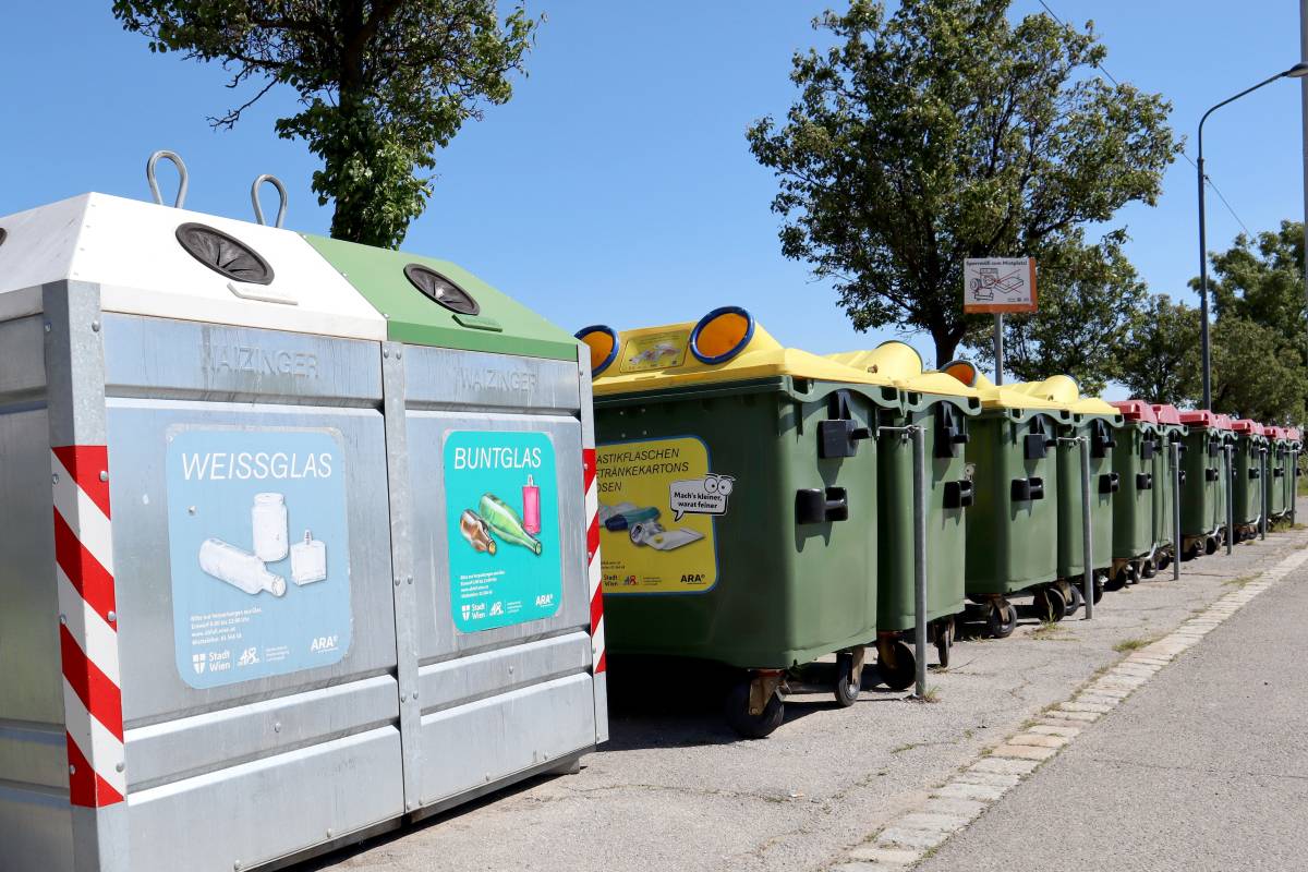 Diverse Müllcontainer zur Mülltrennung entlang einer Straße mit Bäumen und blauem Himmel im Hintergrund.