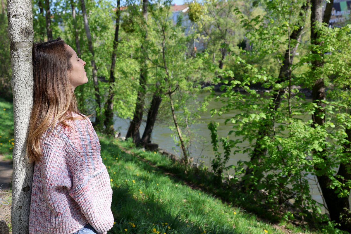 Frau lehnt an Baum an einem Fluß umgeben von Bäumen.